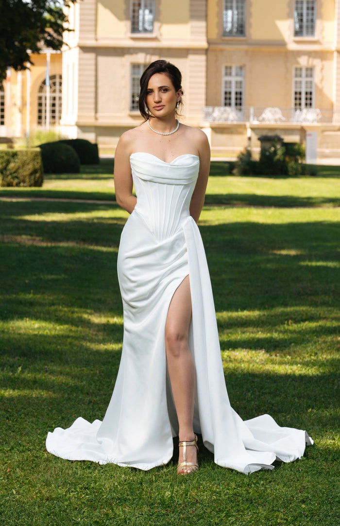 Robe de mariée OCTAVIA en satin avec corset baleiné, drapé et fente haute, vue de face devant un château